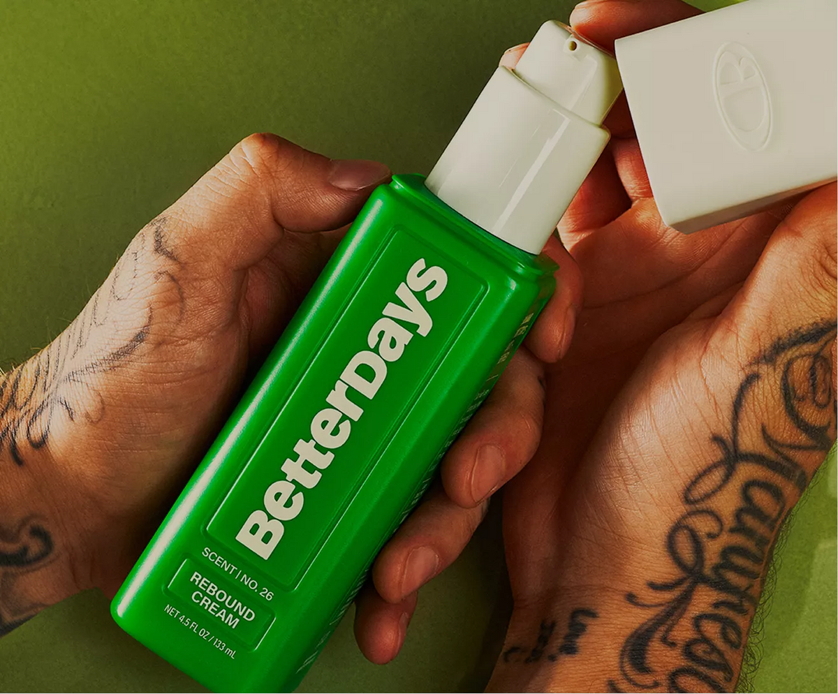 Vic's hands holding a bright green BetterDays Rebound Cream bottle with a white pump, while another hand holds the white cap against an olive green background.